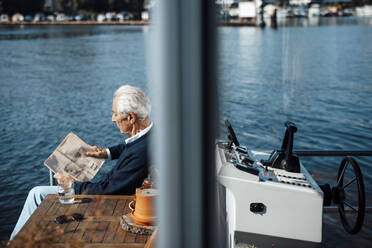 Senior man reading newspaper at houseboat on sunny day - GUSF06858