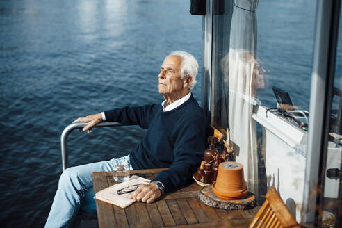 Senior man sitting by table at houseboat on sunny day - GUSF06860