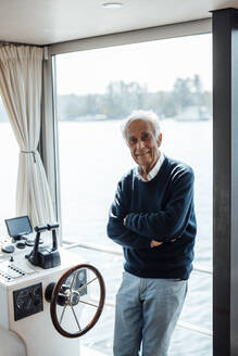 Lächelnder älterer Mann mit verschränkten Armen, der am Fenster eines Hausbootes lehnt - GUSF06877