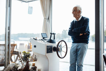 Älterer Mann steht mit verschränkten Armen am Fenster eines Hausbootes - GUSF06878