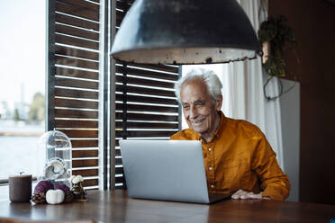 Smiling senior man with laptop sitting at table in houseboat - GUSF06882