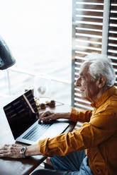 Thoughtful senior man with laptop sitting at table in houseboat - GUSF06883
