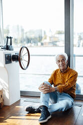 Smiling senior man with white hair sitting by window at houseboat - GUSF06886