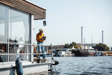 Älterer Mann mit Tablet-PC an der Reling eines Hausbootes an einem sonnigen Tag - GUSF06892