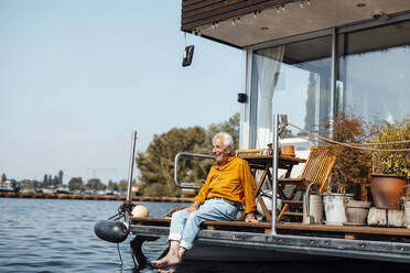 Fröhlicher älterer Mann sitzt auf einem Hausboot an einem sonnigen Tag - GUSF06899