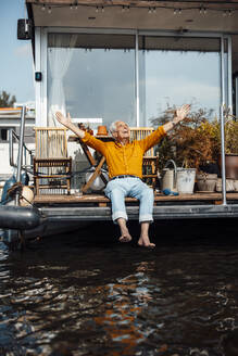Glücklicher älterer Mann mit ausgestreckten Armen, der auf einem Hausboot sitzt und sich an einem sonnigen Tag erfreut - GUSF06902