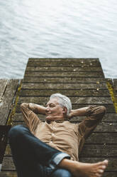 Lächelnder älterer Mann mit Händen hinter dem Kopf auf einem Steg am See liegend - GUSF06913