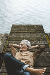 Smiling senior man with hands behind head lying on jetty by lake - GUSF06913