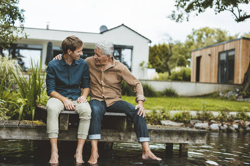 Älterer Mann im Gespräch mit seinem Sohn, der auf einem Steg am See im Hinterhof sitzt - GUSF06915