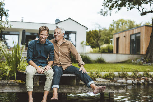 Happy father and son sitting on jetty by lake at backyard - GUSF06916