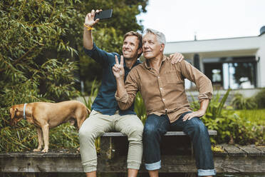 Älterer Mann gestikuliert Friedenszeichen nehmen Selfie mit Sohn durch Handy im Hinterhof - GUSF06921
