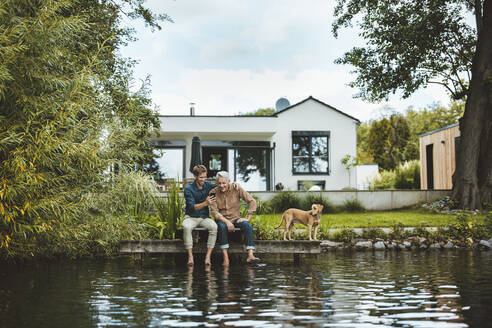 Glücklicher Sohn, der sein Smartphone mit seinem Vater teilt, der auf einem Steg am See im Hinterhof sitzt - GUSF06923