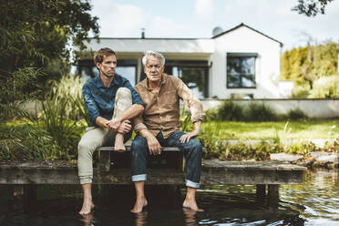 Senior man with son sitting on jetty by lake at backyard - GUSF06924