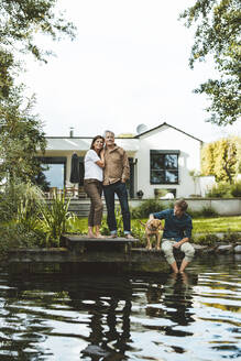 Man with woman standing by son and dog sitting by lake at backyard - GUSF06938