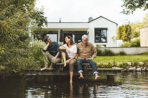 Mother and son talking by senior man sitting on jetty by lake at backyard - GUSF06939