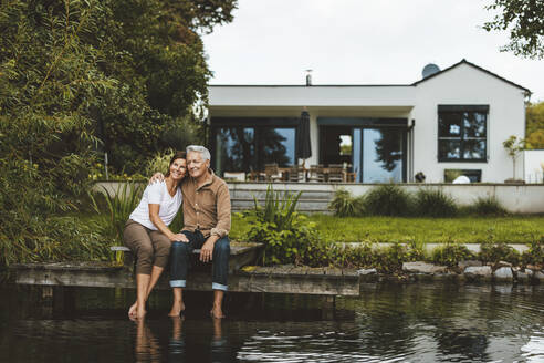 Smiling senior man sitting arm around with woman on jetty by lake - GUSF06943