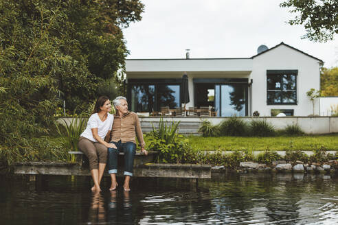 Lächelnde Frau mit Mann sitzt auf Steg am See im Hinterhof - GUSF06944