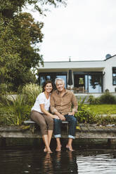 Lächelnder älterer Mann und Frau sitzen auf einem Steg am See im Garten - GUSF06945
