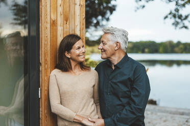 Smiling senior man looking at woman standing by wooden wall - GUSF06971