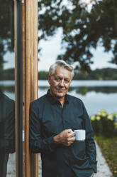 Älterer Mann mit Kaffeetasse an einer Glaswand im Hinterhof stehend - GUSF06974