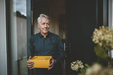 Smiling senior man with white hair holding package at doorway - GUSF07029