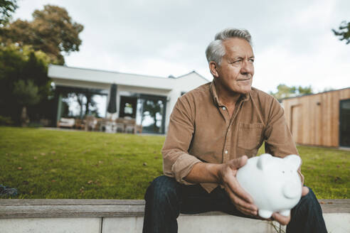 Thoughtful smiling senior man with piggy bank sitting at backyard - GUSF07035