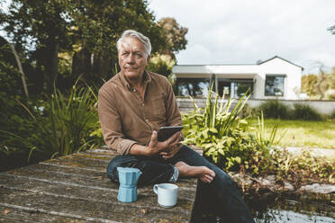 Älterer Mann mit Tablet-PC sitzt auf Steg am See im Hinterhof - GUSF07040