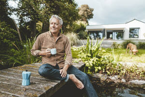 Smiling senior man with coffee cup sitting on jetty by lake at backyard - GUSF07041