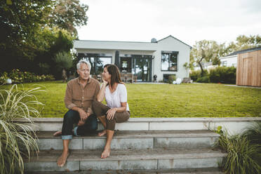 Senior man talking with woman sitting on steps at backyard - GUSF07046