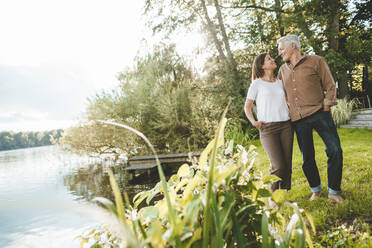 Lächelnder Mann und lächelnde Frau mit den Händen in den Taschen schauen sich am See an - GUSF07050