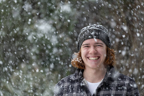 Lächelnder junger Mann mit Strickmütze genießt den Schneefall - LBF03628