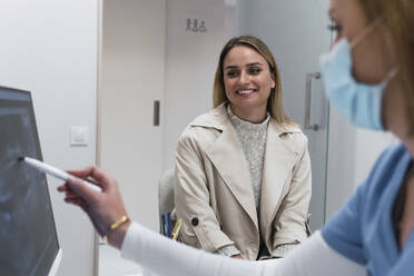 Smiling young woman looking at dentist discussing over intraoral scanner screen at clinic - PNAF03084