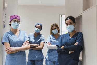 Doctors wearing protective face masks standing with arms crossed in corridor at hospital - PNAF03092