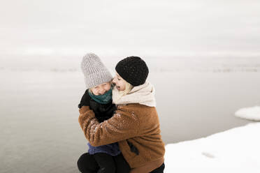 Mother embracing daughter by lake in winter - ANF00082