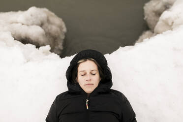 Woman with eyes closed lying down on snow by lake in winter - ANF00098