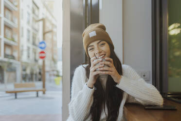Lächelnde Frau mit Strickmütze sitzt in einem Straßencafé - MTBF01163
