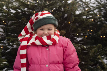 Glückliches Mädchen mit gestreiftem Schal vor einem Weihnachtsbaum stehend - SSGF00519