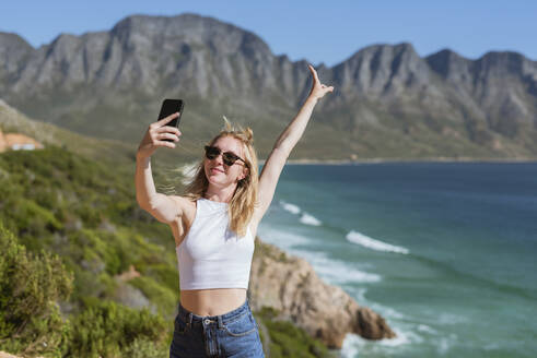 Lächelnde junge Frau mit erhobener Hand, die ein Selfie mit ihrem Mobiltelefon an einem sonnigen Tag macht - MEF00118