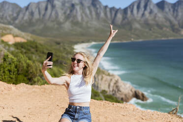 Glückliche Frau gestikuliert Friedenszeichen unter Selfie durch Handy auf sonnigen Tag - MEF00120