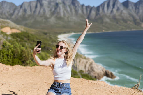 Glückliche Frau gestikuliert Friedenszeichen unter Selfie durch Handy auf sonnigen Tag - MEF00120