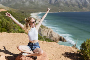 Glückliche junge Frau mit erhobenen Armen sitzt auf einem Felsen und genießt den sonnigen Tag - MEF00121