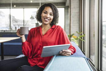 Lächelnde Geschäftsfrau mit Kaffeetasse und Tablet-PC im Büro - UUF25402