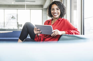Glückliche Geschäftsfrau mit Tablet-PC auf dem Sofa im Büro sitzend - UUF25404