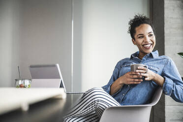 Happy businesswoman with coffee cup sitting on chair in office - UUF25439