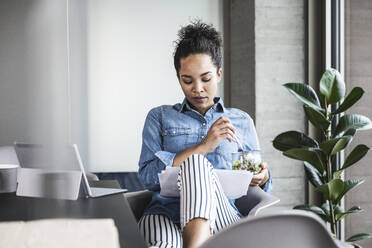 Young businesswoman with food bowl analyzing documents at work place - UUF25440