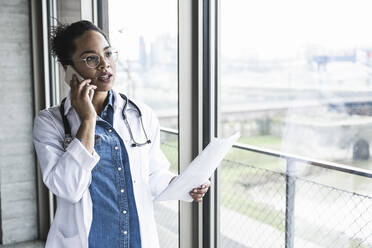 Junger Arzt mit Dokument, der in einer Klinik mit einem Mobiltelefon spricht - UUF25457