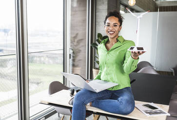 Smiling businesswoman with wind turbine model and laptop in office - UUF25470