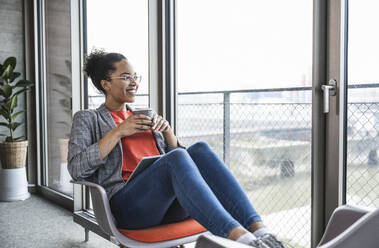 Young businesswoman with coffee cup looking through window in office - UUF25501