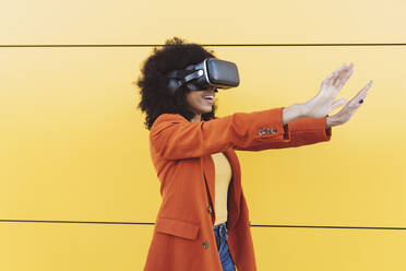 Woman watching through VR glasses by yellow wall - JCCMF05324