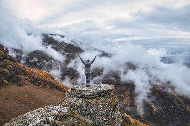 Female hiker standing with raised arms on top of outcrop in autumn mountains of North Caucasus - KNTF06655